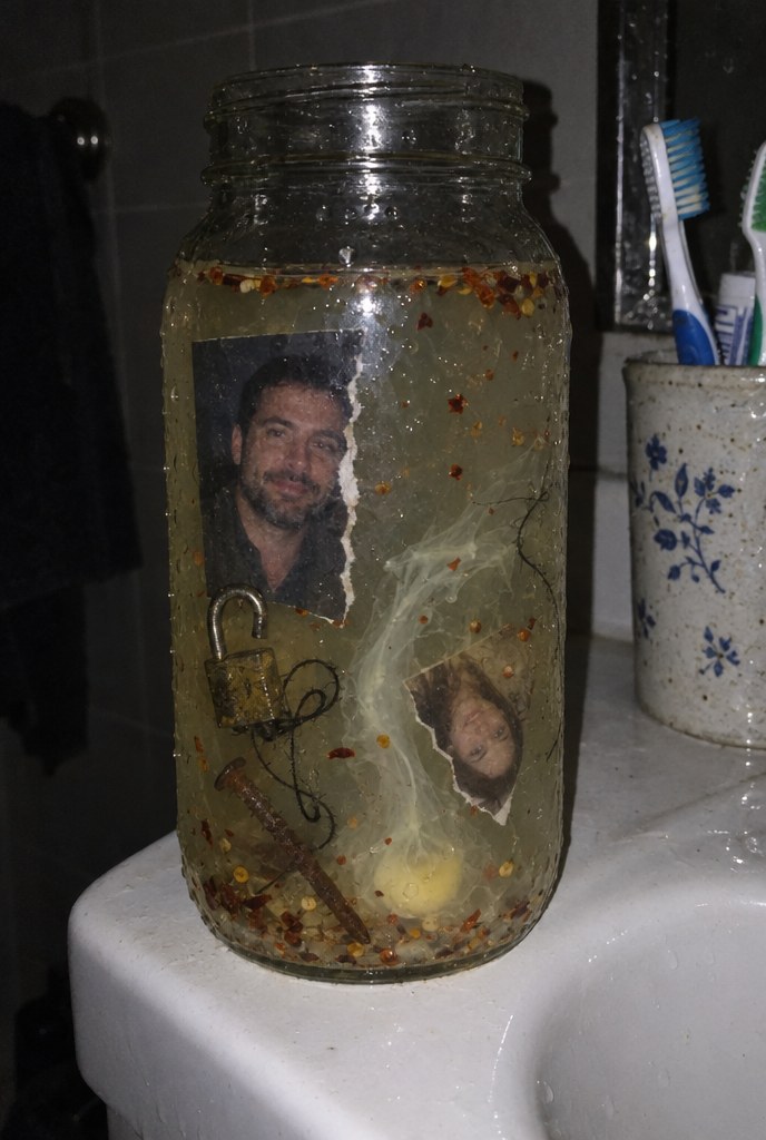 A glass jar on a bathroom sink holding a torn couple photo with the woman's half ripped away, a padlock, a rusty nail and black thread